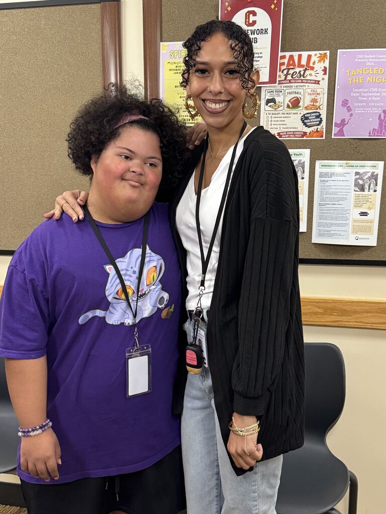 A student and staff member pose together in front of a bulletin board.