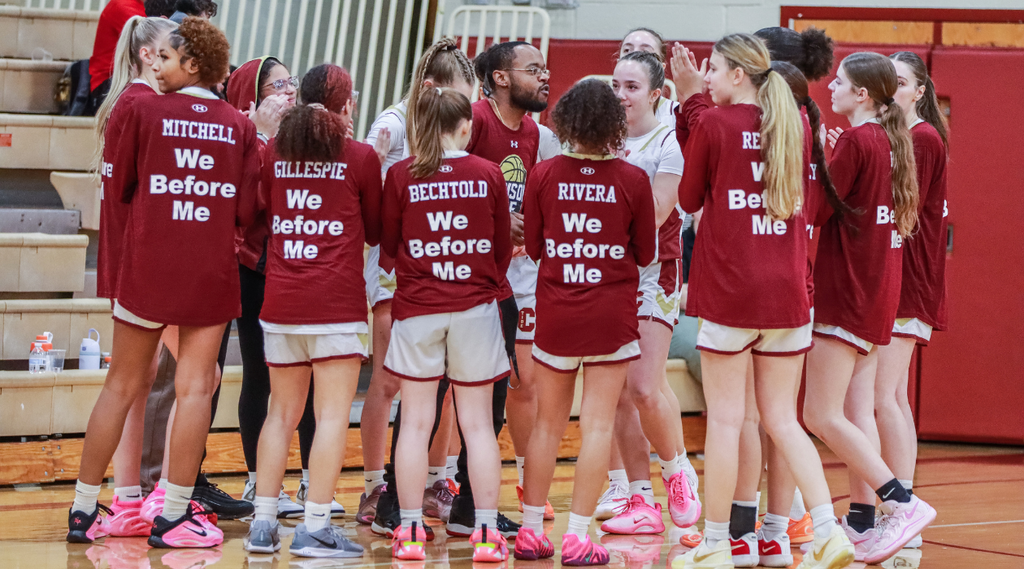 A girls basketball team huddles around  coach keyes in a school gymnasium; players’ warm-up shirts read, “We Before Me.”