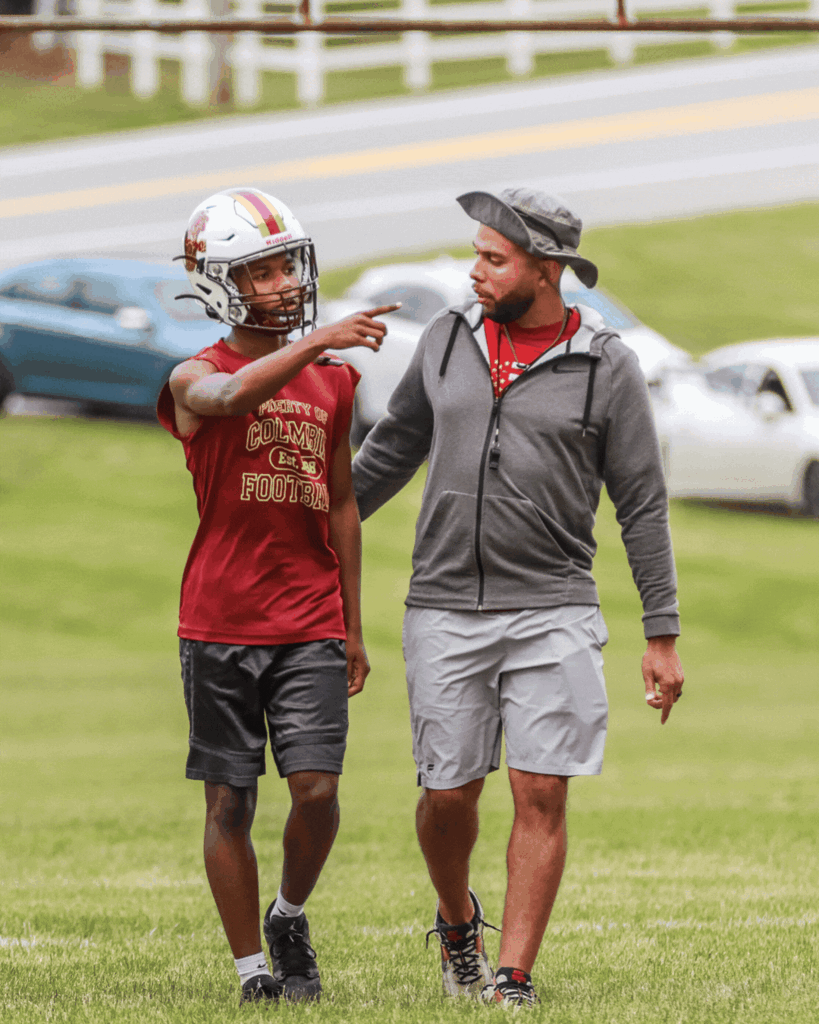 A youth football player in a Columbia football helmet walks across the field beside coach marty moore.