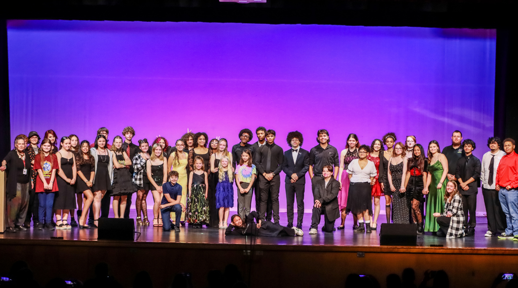 A large group of students and adults pose together on a theater stage under purple lighting.