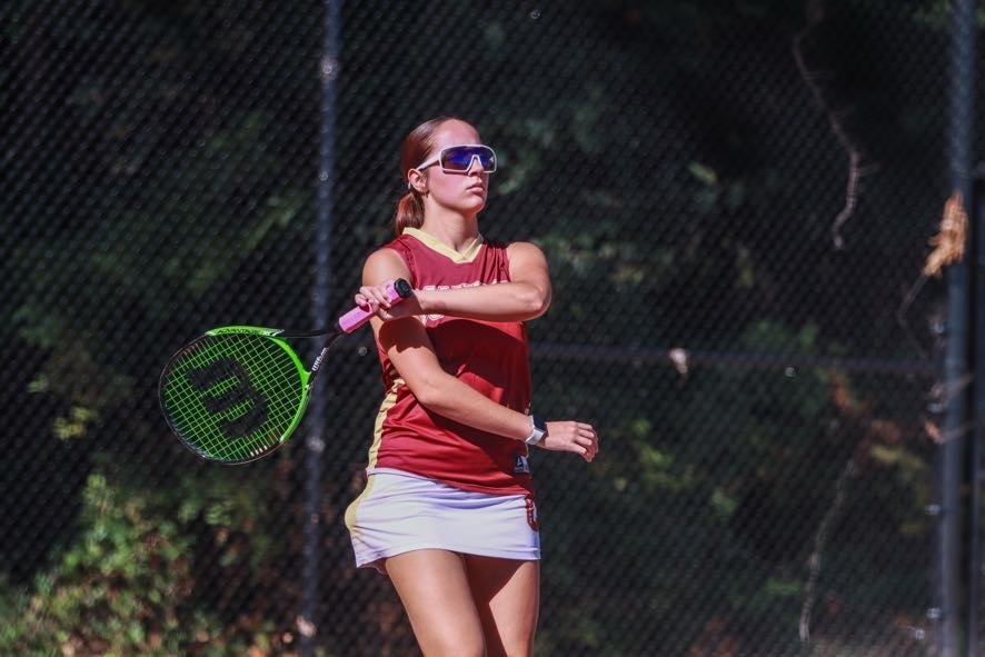 A Columbia tennis player wearing a maroon uniform and sunglasses swings her racket during an outdoor match.