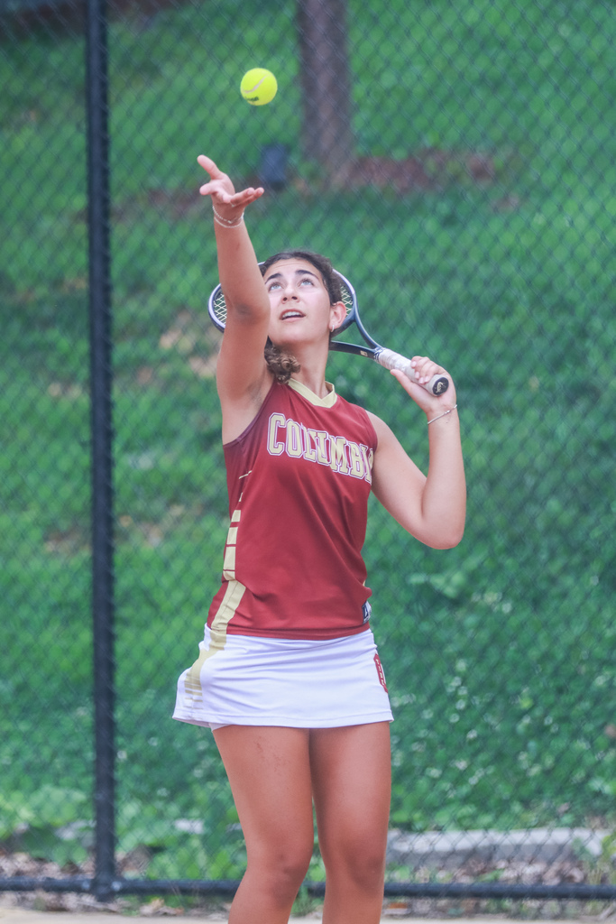A Columbia tennis player tosses a yellow ball into the air while preparing to serve. She wears a maroon Columbia uniform and holds a tennis racket on an outdoor court.