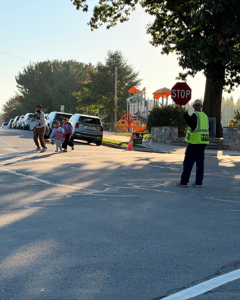 A crossing guard wearing a fluorescent yellow vest holds a red STOP paddle at the edge of a crosswalk while a family with children crosses the street. A playground, parked cars, and long morning shadows are visible.