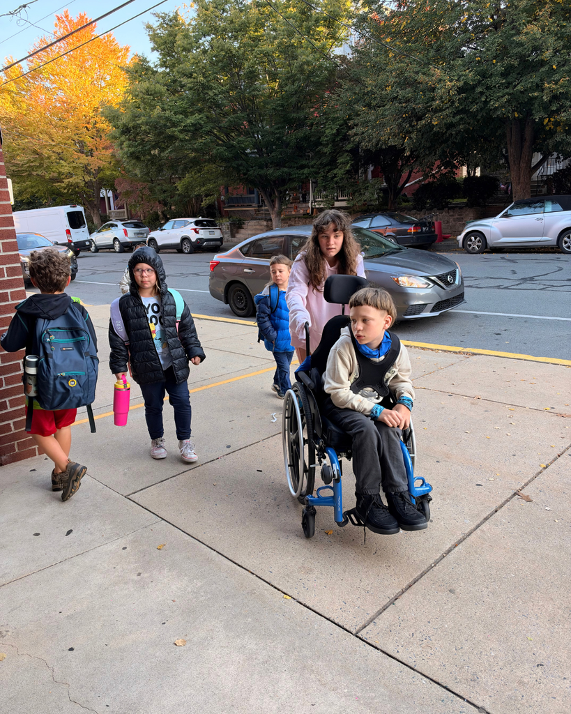 Several students walk along a sidewalk near parked cars and autumn trees. One student uses a blue wheelchair and is pushed by another student; other students walk alongside carrying backpacks and water bottles.