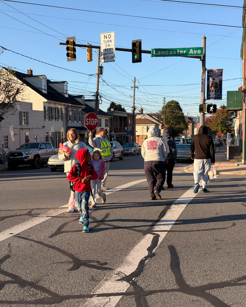 Families and children cross a busy intersection while a crossing guard holds a red STOP paddle. A green street sign reads “Lancaster Ave,” and a white traffic sign reads “NO TURN ON RED 7AM TO 6PM.” The pedestrian signal displays a white walking figure.
