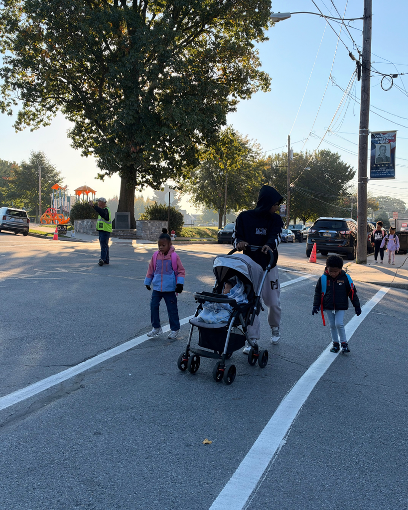 A family with young children crosses a marked crosswalk; one adult pushes a stroller. A crossing guard in a fluorescent yellow vest holds a STOP paddle in the background near a playground and waits for the group to cross safely.