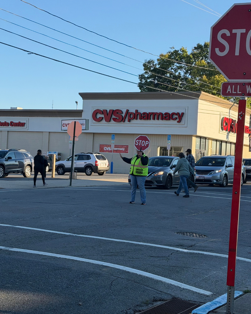 A crossing guard in a fluorescent yellow vest holds a red STOP paddle in the street outside a CVS/pharmacy as pedestrians cross and cars wait nearby. A large stop sign in the foreground reads “STOP” and “ALL WAY.