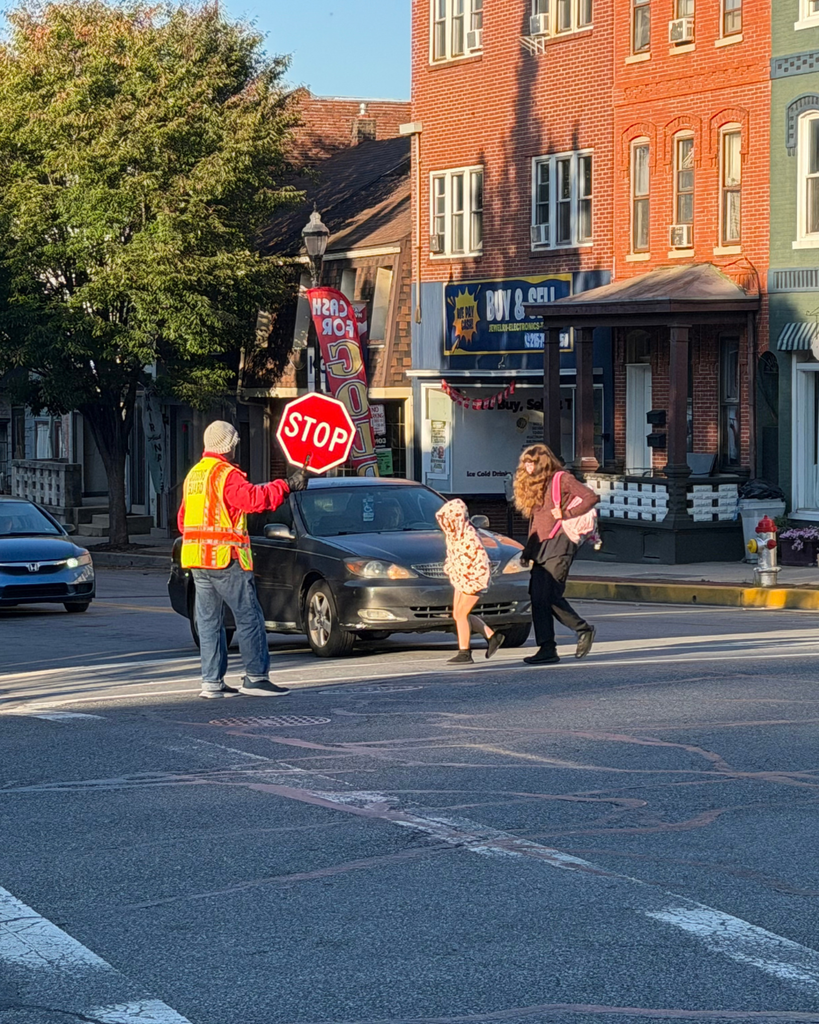 A crossing guard in a fluorescent yellow-and-orange safety vest holds a red STOP paddle while an adult and child cross the street in front of stopped traffic. Brick storefronts line the opposite side of the street; a vertical sign reads “CASH FOR gold.
