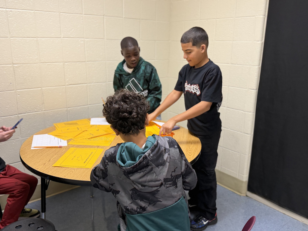 Four students work together at a table on a math activity, using worksheets, pencils, and calculators. The students are smiling and engaged in their work.