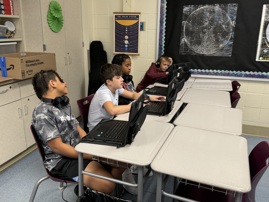 Three students work on laptops at classroom desks while completing an independent learning activity. A solar system display and student artwork decorate the classroom.