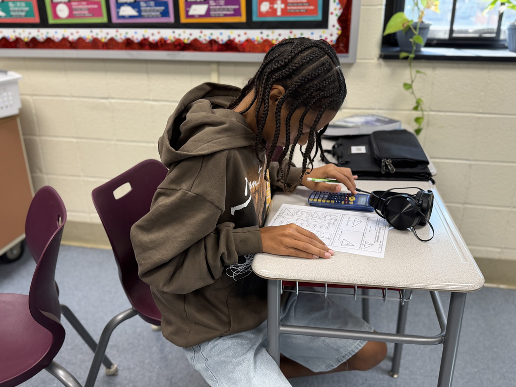 A student works independently on a math worksheet while using a calculator. Headphones rest on the desk beside the student.