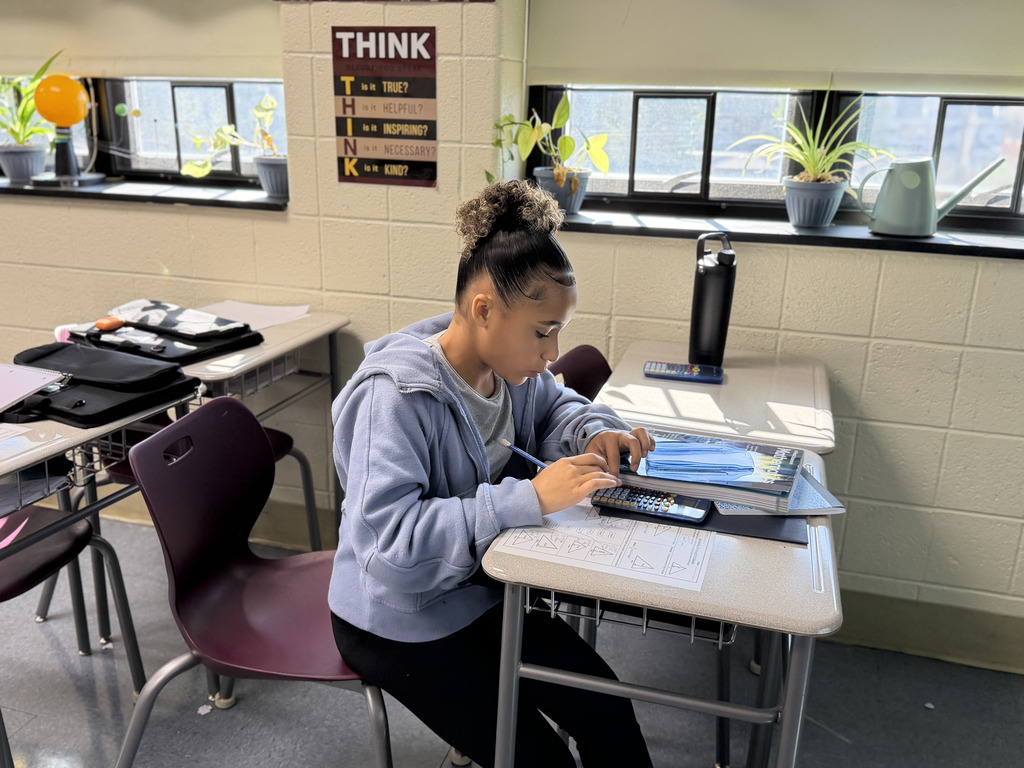 A student works independently on a math worksheet while using a calculator. Headphones rest on the desk beside the student.