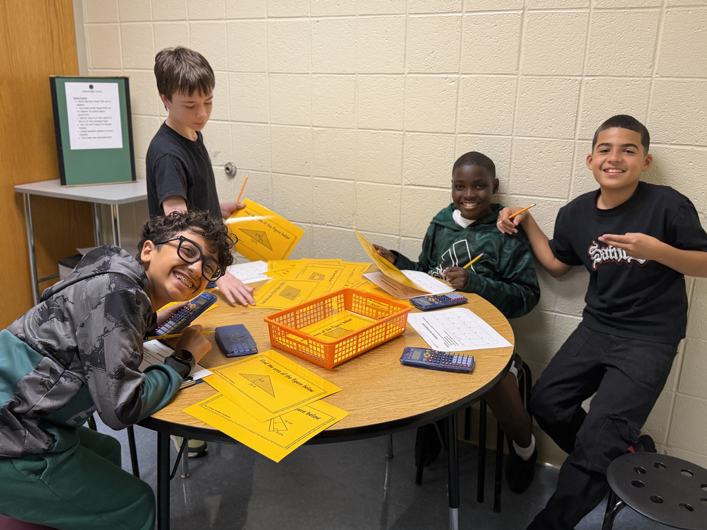 Four students work together at a table on a math activity, using worksheets, pencils, and calculators. The students are smiling and engaged in their work.