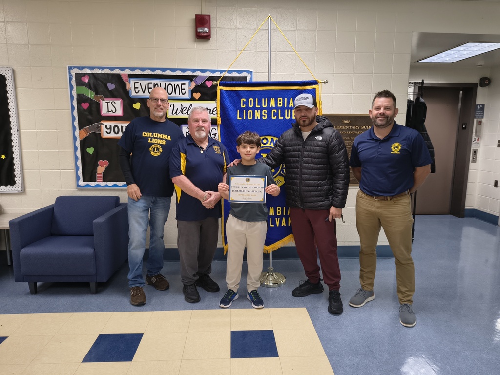 A student stands with four men from the Columbia Lions Club, holding a Student of the Month certificate for September 2026. The group poses in front of a blue and gold Columbia Lions Club banner in the school hallway. A colorful welcome bulletin board and blue chairs are visible in the background.