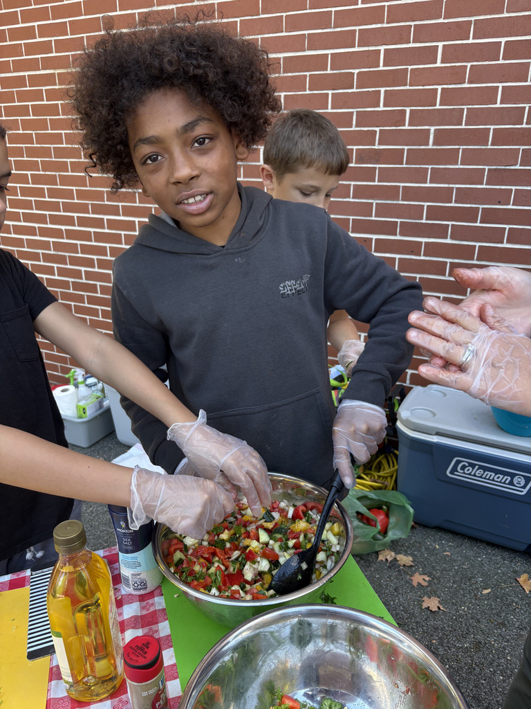 Students wearing gloves work together to mix freshly chopped vegetables in a large metal bowl while preparing Israeli salad outdoors.