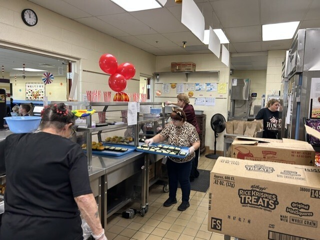 Food service staff members prepare lunch trays in a school cafeteria kitchen decorated with red balloons and carnival-themed signs. A large box of Rice Krispies Treats is visible in the foreground.