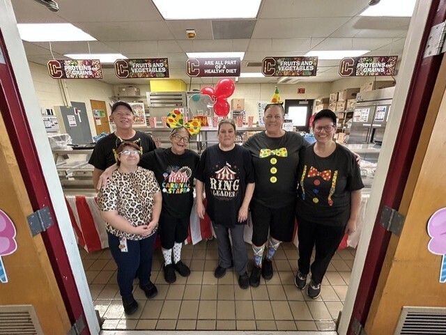 Six school food service staff members pose together in the cafeteria kitchen wearing carnival-themed clothing. Colorful carnival decorations and banners about healthy food groups hang behind them.