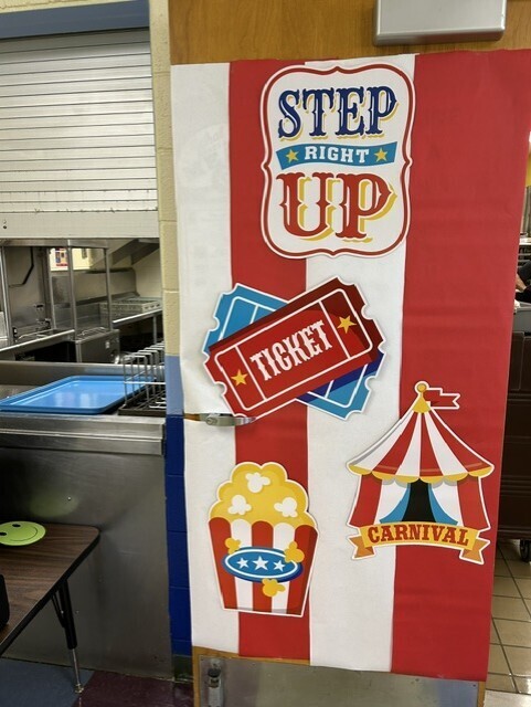 Red, white, and blue carnival-themed sign at the cafeteria entrance. Text reads: “STEP RIGHT UP,” “TICKET,” and “CARNIVAL,” with illustrations of tickets, popcorn, and a circus tent.