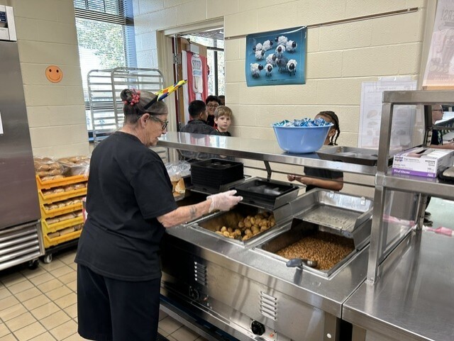 A cafeteria staff member wearing a colorful headband and gloves serves lunch from a hot-food station while students wait on the other side of the counter.