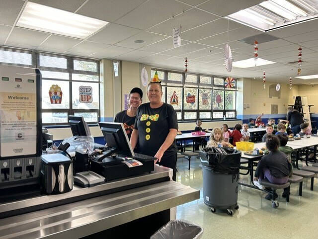 Two school food service staff members in carnival-themed shirts smile behind a cafeteria checkout counter while elementary students eat lunch at tables decorated with hanging red, white, and blue carnival decorations.