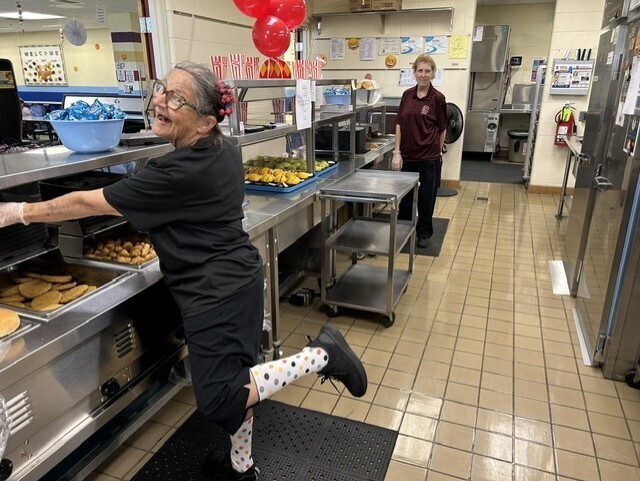 A smiling cafeteria staff member in a carnival-themed outfit poses playfully beside trays of lunch food, while another staff member stands farther back in the kitchen. Red balloons and carnival decorations are visible.