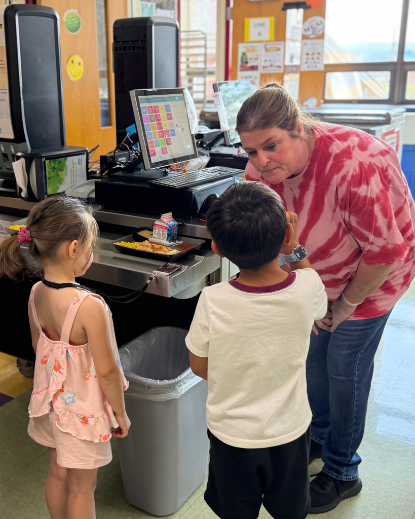 A cafeteria staff member wearing a pink tie-dye shirt leans down to speak with two young students at a school lunch checkout station. A meal tray with milk and food sits on the counter beside a point-of-sale screen.