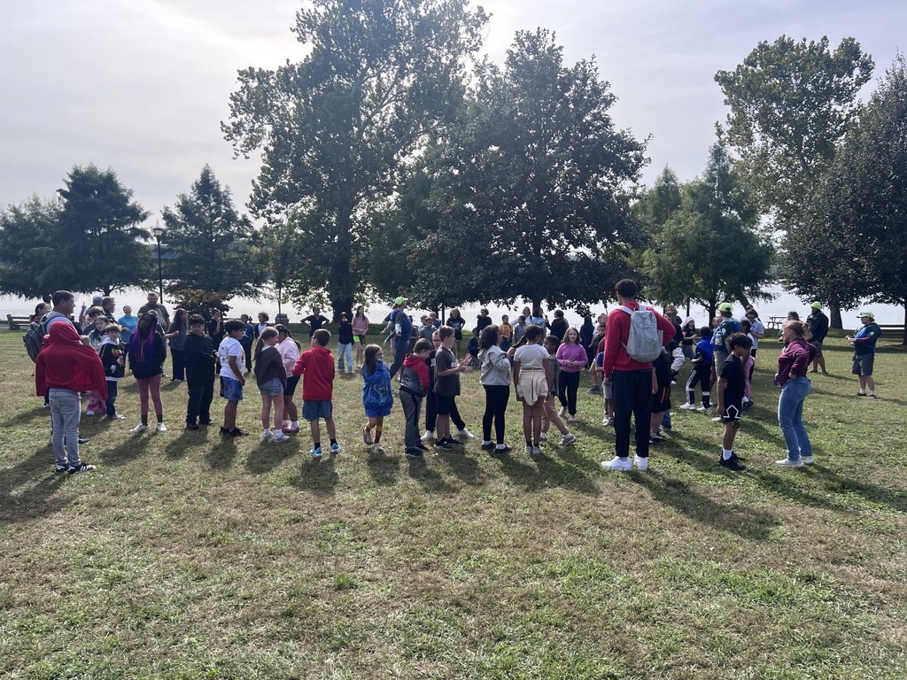 A large group of students and chaperones gathers on a grassy riverside area beneath trees during an outdoor educational field trip.