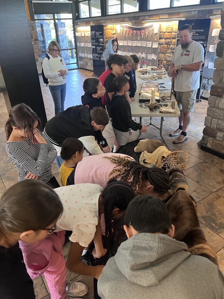 Students gather around a table indoors to learn about rain gardens, examining educational displays, plant samples, and water-related materials with an instructor.