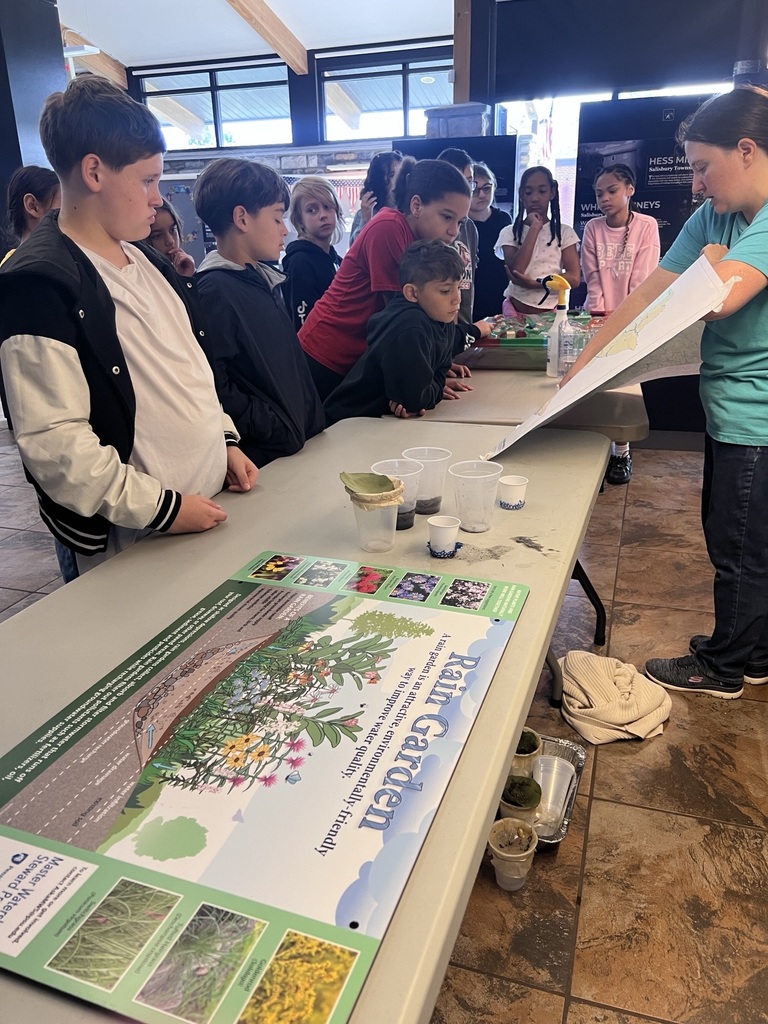 Students gather around a table indoors to learn about rain gardens, examining educational displays, plant samples, and water-related materials with an instructor.