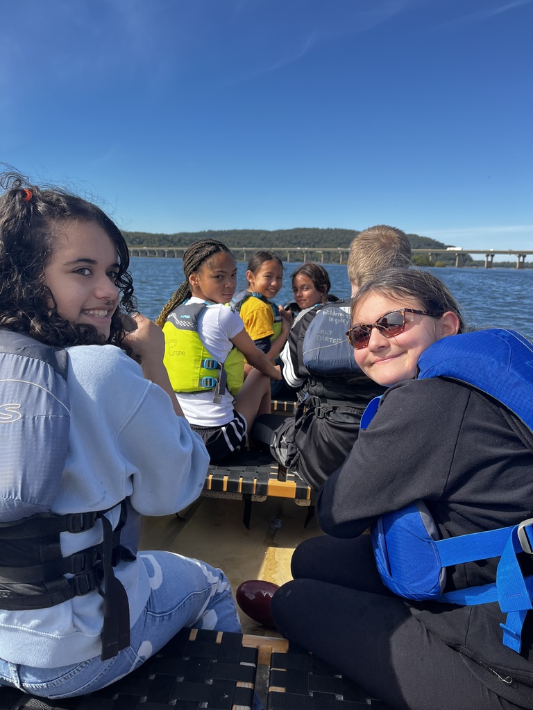 Students wearing life jackets sit together in a canoe on the river, smiling toward the camera as they participate in an outdoor water adventure.