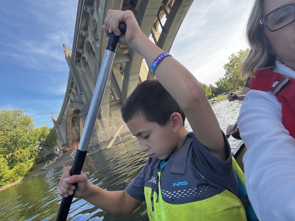 A student wearing a life jacket practices paddling a canoe on the river beneath a large bridge, with an adult nearby providing guidance.