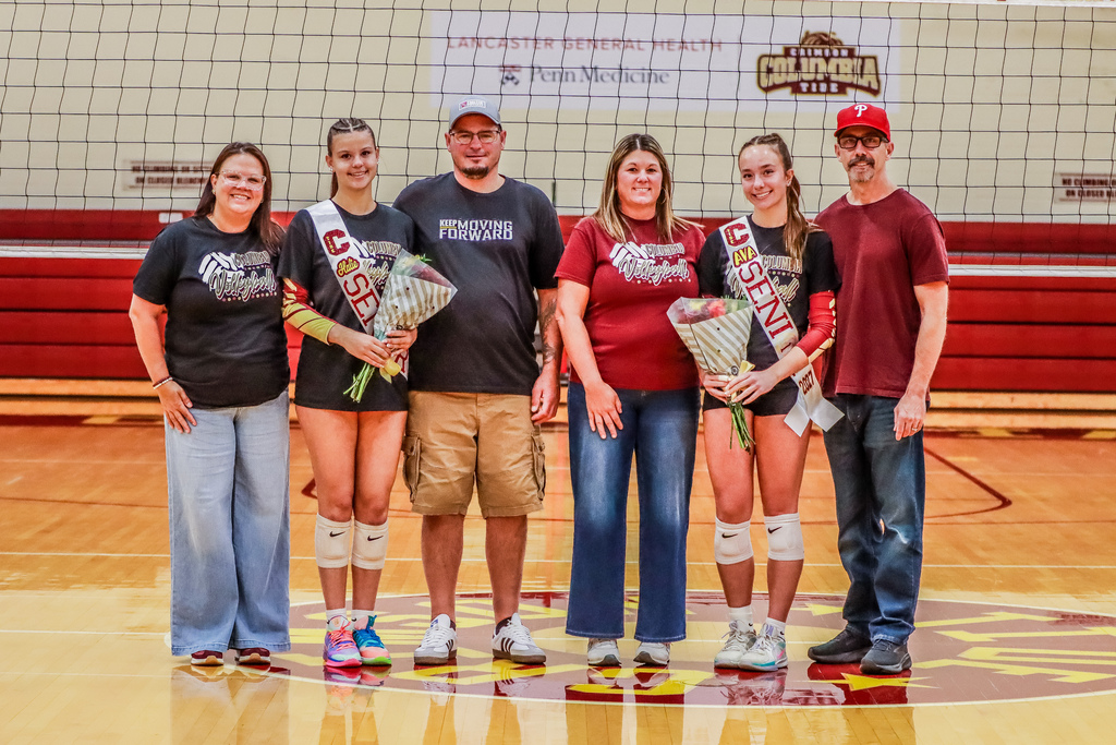 Columbia volleyball seniors Halie Rineer and Ava Broome stand with family members on the gym court during Senior Night. Both wear “Senior” sashes and hold bouquets in front of the volleyball net.