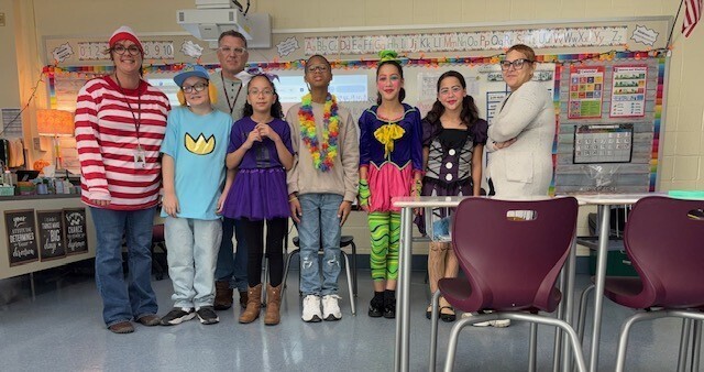 A group of students and staff pose together in a classroom wearing costumes inspired by storybook and popular characters, including a red-and-white striped shirt and hat, colorful outfits, and a black-and-white polka-dot dress.