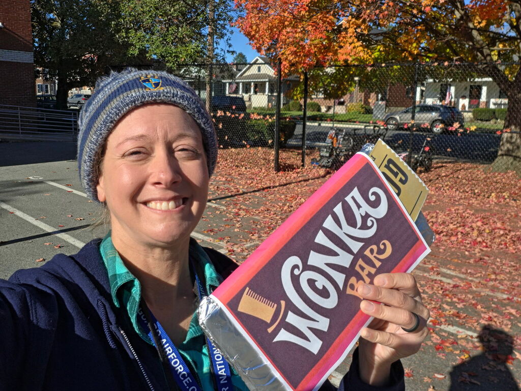 A smiling staff member stands outside among colorful autumn leaves, holding a sign that says “Magic Book” during Storybook Character Day.