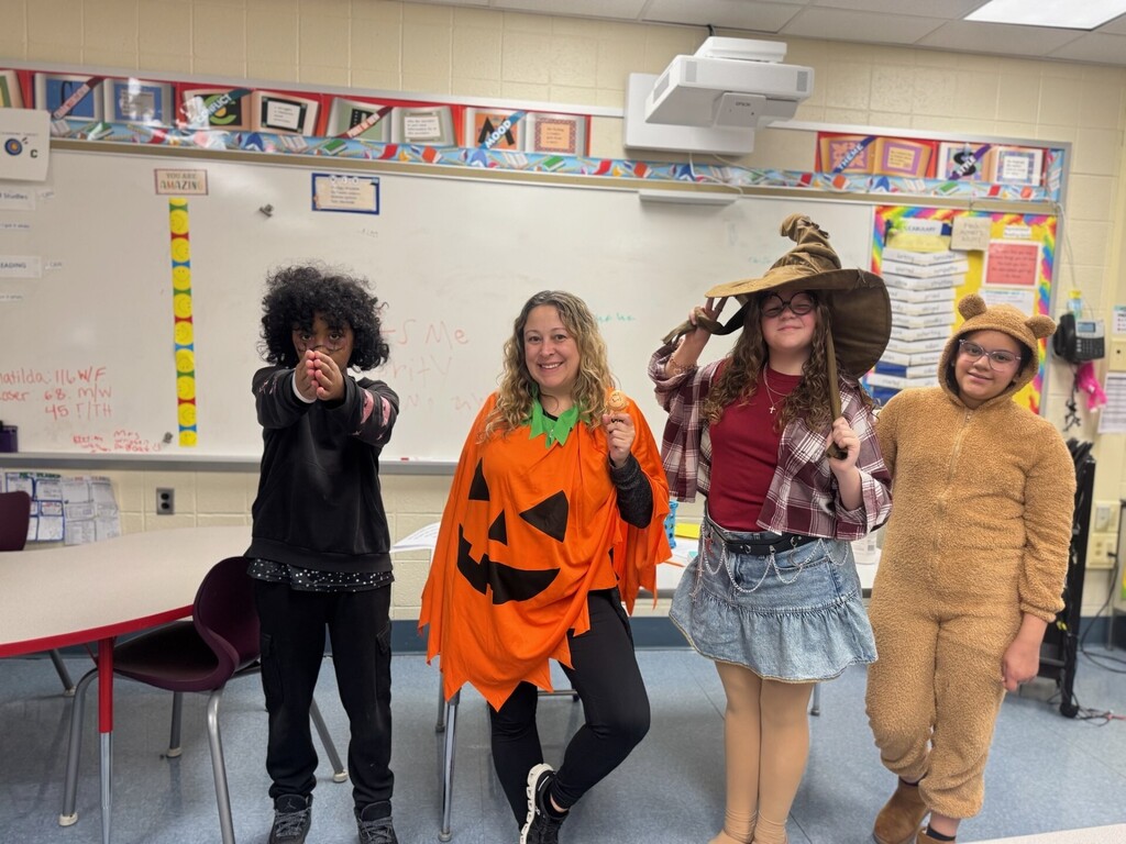 A staff member dressed as a jack-o’-lantern poses with three students dressed in costumes, including a black outfit with curly hair, a wizard with a large hat, and a brown bear costume, inside a classroom.