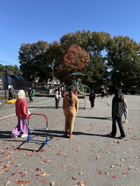 Children and adults gather in a sunny school playground, moving among wheeled frames beneath colorful autumn trees; the scene feels calm and active.