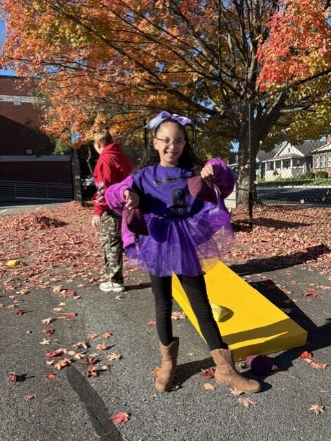 A smiling child in a purple witch costume poses beside a yellow slide beneath colorful autumn trees, with fallen leaves covering the playground.