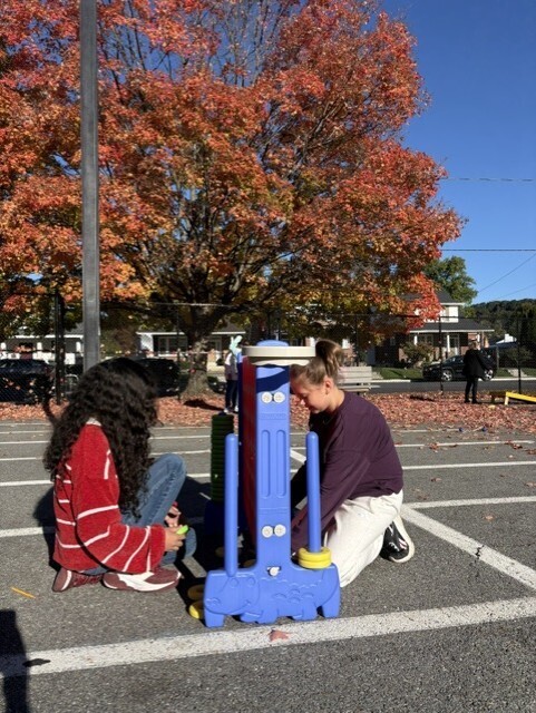 Two people crouch on a school playground, operating a blue hand-pump toy as autumn leaves glow overhead in the bright sun, creating a playful mood.