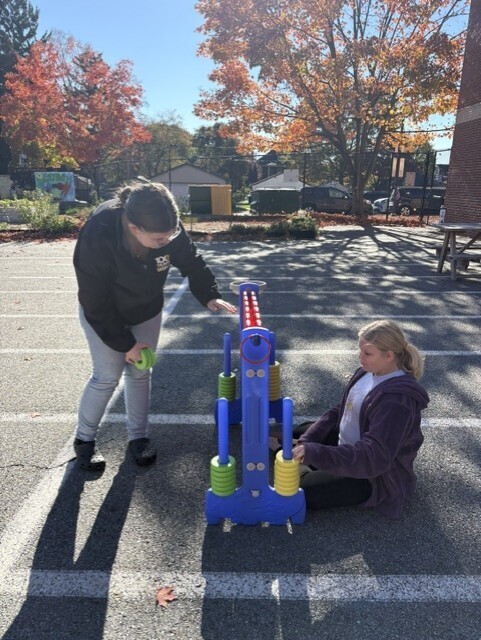 Two people play a sidewalk ring-toss game in a sunny parking lot, with autumn trees behind them; the scene feels casual and cheerful.