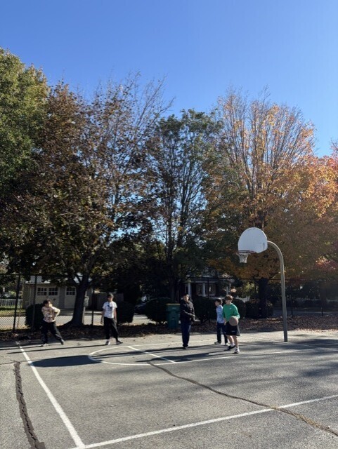 Children play basketball on an outdoor court beneath autumn trees, with a hoop, fallen leaves, and clear blue sky creating a bright, relaxed neighborhood scene.