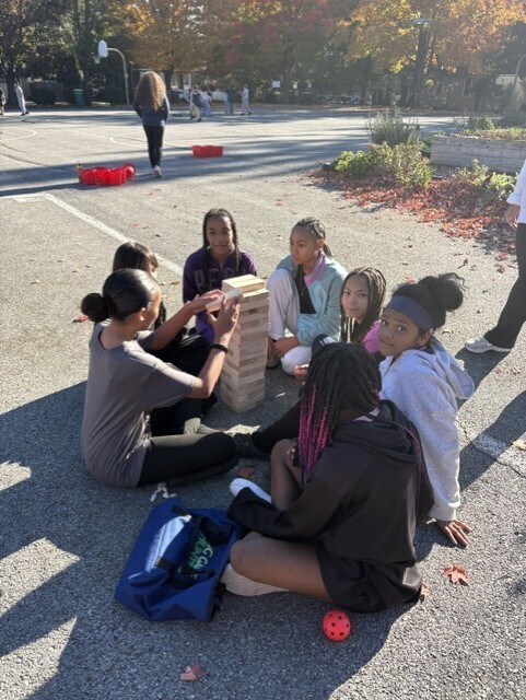 A group of schoolgirls sits in a sunny autumn parking lot, working together to build a tall tower from wooden blocks; a relaxed, collaborative outdoor activity.