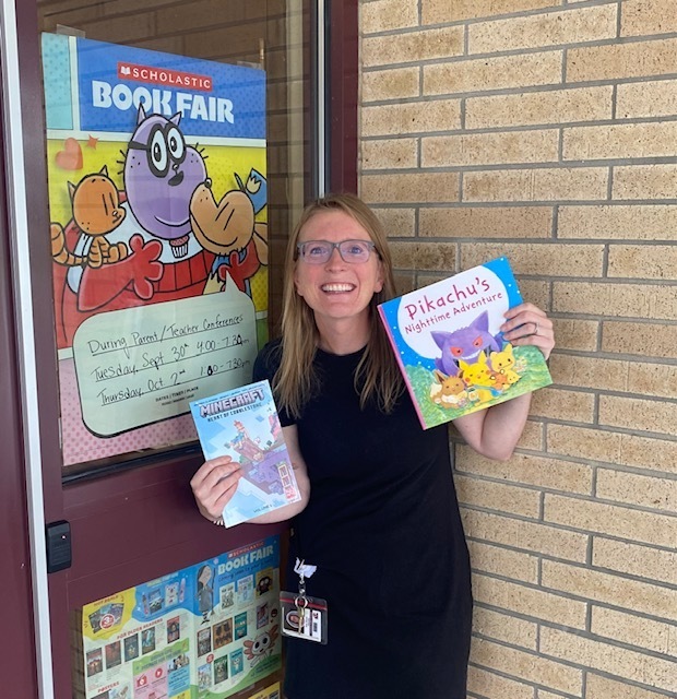 Mrs. Moore is standing by the book fair poster and holding two books that will be at the book fair.