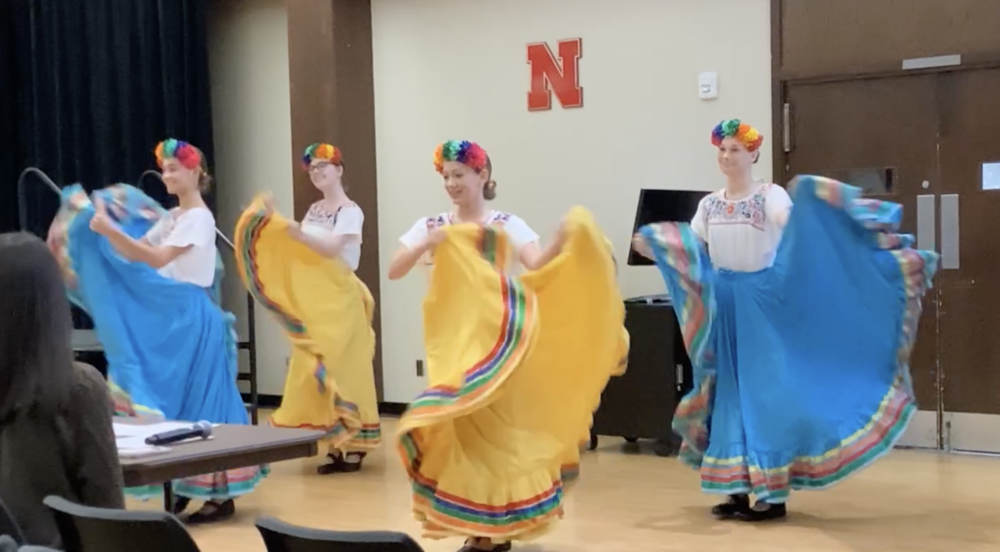 4 high school females wearing traditional mexican attire competing in a dance routine,  Two skirts are blue with rainbow trim and 2 skirts are yellow with rainbow trim.  they are all wearing flower headpieces that are rainbow colored.