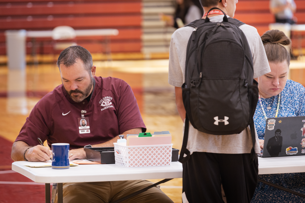 A pair of staff help a freshmen approve his schedule. The student is a table, and has his back to the camera.