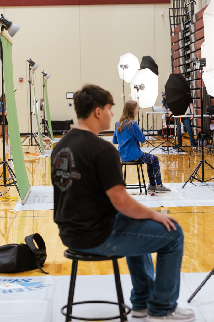 Two sophomores pose for student pictures. They are on stools in front with a green screen and flashes set around them.