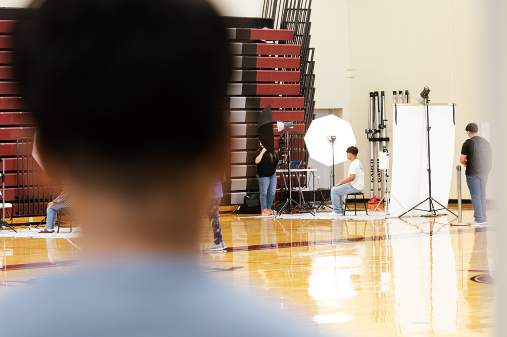 A CHS sophomore watches a classmate take his class photo. He is in the foreground and blurry and the student is in the background, in focus and set in front of flashes.