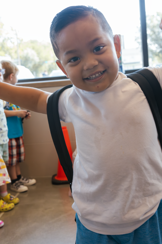 A preschool boy smiles close to the camera as he waits for his first day of school in the vestibule at Kramer Preschool