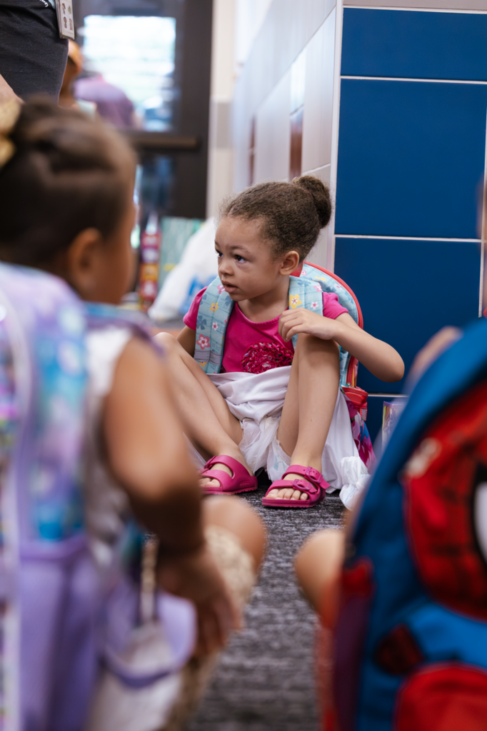 A Kramer preschool student sits in the hallway awaiting her first day of school. Two other students are in the foreground, out of focus.
