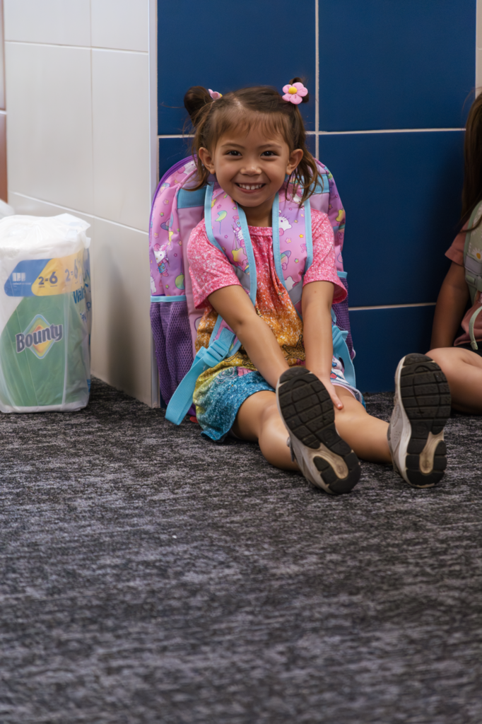 A Kramer preschool student sits in the hallway awaiting the first day of school. She is smiling, wearing a rainbow shirt and colorful backpack.