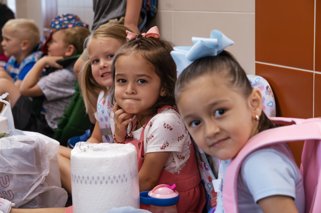 Three Kremer preschool girls smile while sitting in line outside their classroom.